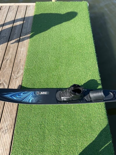 The image depicts a single black "OMNI" brand water ski resting on a strip of green artificial turf, positioned at the edge of a body of water in Coruche, Portugal. The water ski features a blue and white swirling graphic design on its surface and is equipped with a black boot-style front binding with laces and a black open-toe rear binding. To the left of the artificial turf is a weathered wooden dock composed of parallel planks. A dark, calm body of water is visible along the right edge of the artificial turf and beyond the wooden dock. The scene is illuminated by strong sunlight, casting a prominent human shadow from the upper left corner across the wooden dock and a portion of the artificial grass. Another, less defined shadow is also present on the artificial turf. No individuals are directly visible.