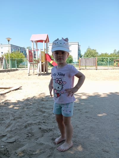 A young girl stands barefoot in a sand-covered playground in Radzymin, Poland. She is wearing a light pink t-shirt with a "Hello Kitty" graphic, light blue shorts with a bunny print, and a white baseball cap with bunny ears. Behind her, a red and yellow playground structure is visible. The background shows apartment buildings and trees under a clear blue sky. The sun casts shadows on the sand.
