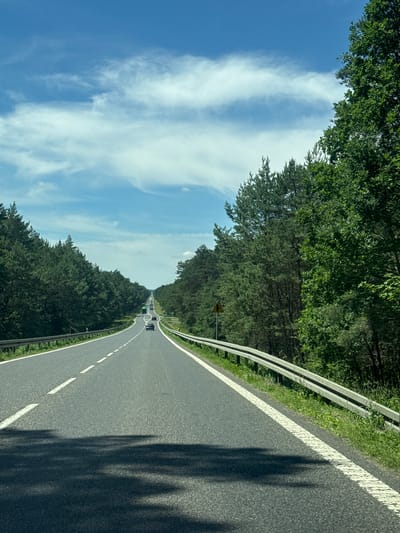 The image shows a view from a vehicle driving on a paved road lined with a metal guardrail and dense green trees on both sides. The road has a white dashed line marking the lanes. The sky above is blue with scattered white clouds, suggesting a clear day. In the distance, several cars are visible on the road, indicating traffic. There are no people clearly visible in the image, but shadows suggest their presence within the vehicle. The scene appears to be a scenic drive through a wooded area. A yellow triangular road sign with a symbol is visible on the right side of the road. The lighting suggests it is daytime, likely morning or afternoon. The overall mood is peaceful and natural. The location is identified as Popowo-Letnisko, Poland.