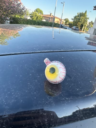 The image shows a close-up view of a novelty candy shaped like an eyeball, resting on the dusty, dark surface of a car hood. The candy has a white outer part with red veins, a yellow iris, and a dark green pupil. A small red and white sticker with a symbol of a child with a line through it is visible on the packaging.
The background is a reflection of an outdoor scene with a bright blue sky, suggesting it is daytime with clear weather. Trees with green foliage and some with pink blossoms, as well as buildings and streetlights, are visible in the reflection. The overall impression is that the photo was taken outdoors on a sunny day, possibly in a residential or semi-urban area. The dust on the car hood suggests the vehicle has been parked outside.
The image is oriented vertically, with the car hood occupying the majority of the frame. The reflection creates a mirror image of the environment, adding depth and context to the scene. No people are directly visible, but their presence is implied by the car and the surrounding environment. The candy object is the primary focus, with its playful and slightly unsettling design.
