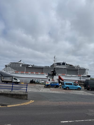 A large white cruise ship, identified by the "MSC" logo, is docked at a port under a cloudy sky. The ship, named "Virtuosa" on its side, is positioned parallel to the quay. In the foreground, a paved road with double yellow lines runs horizontally. To the left of the road, a blue metal railing separates the walkway from the port area. Several vehicles are visible on the quay, including a white truck with a flatbed, a blue car, a light blue van, and a dark colored van. A red crane is attached to a structure on the ship. The environment appears to be a commercial port. The location is the port of Lerwick, United Kingdom, which is a hub for cruise ships and cargo vessels in the Shetland Islands.