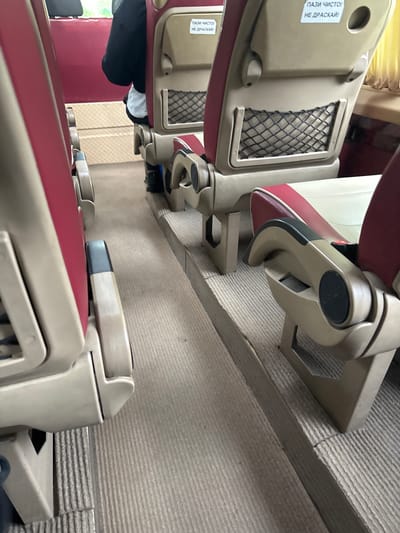 The image shows the interior of a train carriage. Red and beige upholstered seats are visible, arranged in rows. There is a mesh net on the back of some seats and a sign in Bulgarian "ПАЗИ ЧИСТО! НЕ ДРАСКАЙ" meaning "KEEP CLEAN! DO NOT SCRATCH!". A person's legs, wearing black trousers and white socks, are visible between the seats. The floor of the carriage is carpeted in a gray textured material. The location is Pevets, Bulgaria.