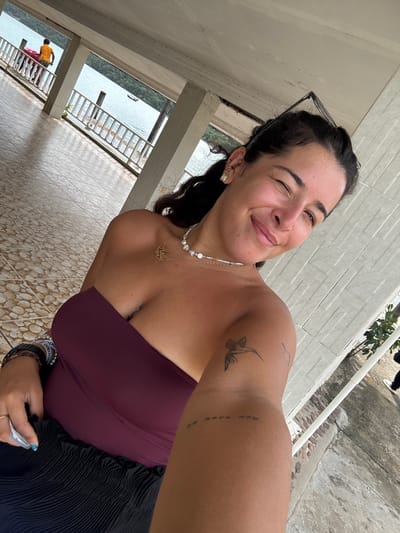 A woman with dark hair is taking a selfie outdoors. She is wearing a strapless plum-colored top and a black skirt. She has several bracelets on her left wrist and a necklace around her neck. She has tattoos on her left arm and thigh. She is winking and smiling at the camera. In the background, there is a railing overlooking a body of water with a boat in the distance. There is also a person wearing a yellow shirt in the background. The flooring is tiled with a pebble pattern. The architecture suggests a covered walkway or balcony, possibly part of a resort or public building. The overall environment suggests a tropical or coastal location. The presence of a person wearing a yellow shirt and the specific tile pattern might be indicative of a location in Preah Sihanouk, Cambodia, though further context would be needed for definitive identification.