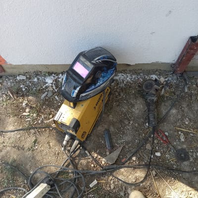 An overhead, high-angle shot captures a welding setup on a dirt and gravel surface in front of a textured white wall. A yellow inverter welding machine is positioned in the center, with a black and blue welding mask placed on top of it. To the right of the welding machine, an angle grinder with a cutting disc is visible, alongside a pair of red-handled pliers. Various electrical cords and a black power strip are scattered on the ground. The environment suggests an outdoor construction or repair site, with debris and dry grass present. The scene is located in Çatalca, Turkey.