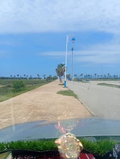 This image captures an outdoor scene, likely along a coastal road or promenade in Martil, Morocco, viewed partially through a reflective surface or window.
The upper portion of the frame is dominated by a clear blue sky with scattered wispy white clouds. Below the sky, a line of identical palm trees and modern streetlights extends into the distance along a paved road. In the left foreground, a distinct white streetlight with ornate blue decorative elements is prominent, alongside other poles and lush green vegetation.
A wide, light-colored paved pathway runs parallel to a grassy verge, which in turn borders an asphalt road. The road has visible white markings. The repetitive planting of palm trees and streetlights suggests a planned urban or coastal landscape.
The right side of the image is partially obscured by what appears to be the interior of a vehicle or a window pane. This section shows a clear, reflective surface. Within this area, there's a patch of vibrant artificial green grass, a hint of a patterned red and black fabric (possibly a rug or cushion), and a decorative, iridescent, rose-shaped object that reflects light with various hues.
The scene is brightly lit, indicating a sunny day, likely morning or early afternoon. No people or visible text are present. The overall impression is one of a well-maintained, bright, and somewhat serene environment.