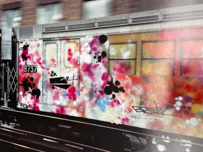 The image shows a subway car in the city of New York, United States. The car is painted with colorful graffiti art. The car is parked at a station. The car is a standard New York subway car, with a number of windows and doors. The car is also marked with the number 8737. The graffiti art is painted in various colors and patterns, some of which are obscured by the blur of motion. The art includes black splatter, pink and white streaks, and an abstract design reminiscent of a ship. The car is parked on a dark black track. The environment is urban and industrial.