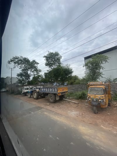 The image depicts a street scene in Bethamcherla, India, under an overcast sky. On the right side of the road, a yellow three-wheeled auto-rickshaw is parked. Adjacent to it, a blue and yellow tractor with a trailer is stopped. To the left of the tractor, a white SUV is parked. In the background, a long, low building with a dark roof is visible, likely an industrial or commercial structure. Trees are scattered throughout the scene, with denser foliage behind a low stone wall separating the road from the background structures. Power lines are strung across the sky. The road itself is paved, with a dirt and gravel area alongside where the vehicles are parked. The overall impression is of a functional, working area within the town.