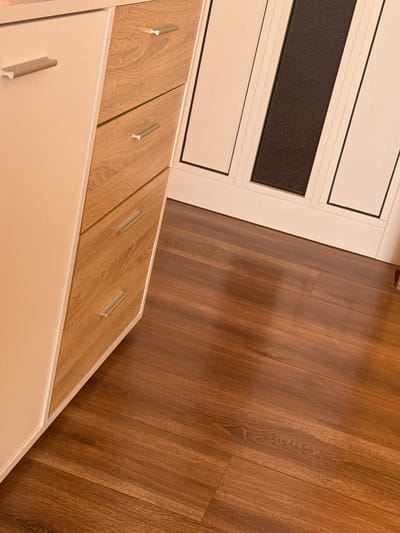 The image shows a partial view of furniture and flooring. A light-colored dresser with wooden-look drawer fronts is positioned at the bottom of the frame. Each drawer has a simple, elongated handle. Above the dresser is a door or wall panel with a white frame and rectangular insets, including a black panel. The floor is made of wooden planks with a brown finish. The location, Rakovski, Bulgaria, cannot be confirmed from the visual elements. No people, actions, or interactions are visible.