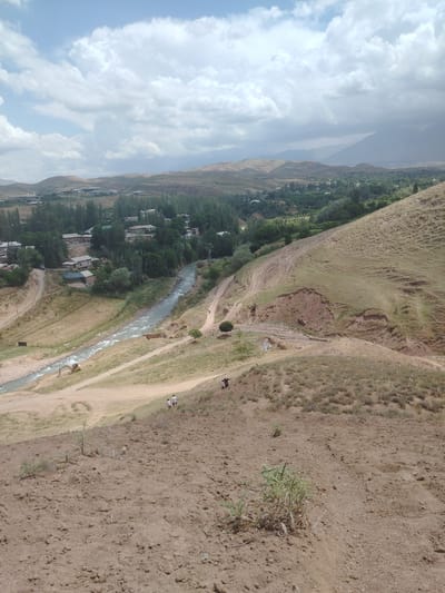 The image captures a rural landscape with a meandering river flowing through a valley in Tashbulak, Uzbekistan. In the foreground, a barren, reddish-brown hillside dominates, with sparse, dry vegetation. Further down the slope, a sandy path winds its way towards the river.

The middle ground reveals a verdant valley with a cluster of houses nestled amongst tall, green trees. A river, characterized by its light-colored, turbulent water, cuts through the valley floor. An excavator, yellow in color, is visible near the riverbank, suggesting ongoing construction or maintenance work. A dirt road runs parallel to the river.

Several individuals are present: two people are walking up the hillside in the middle distance, and a smaller group of three individuals, two adults and a child, are walking down the hillside closer to the foreground.

The background showcases rolling hills that gradually recede into the distance, topped by a hazy, mountainous horizon under a partly cloudy sky. The overall scene depicts a serene, natural environment integrated with a small settlement, characteristic of rural Uzbekistan.
