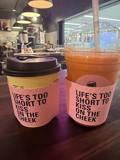 Two drinks, a coffee cup and a large plastic cup with a straw, sit on a dark, reflective surface. The coffee cup is yellow with a dark lid and a pink sleeve that reads "LIFE'S TOO SHORT TO KISS ON THE CHEEK". The plastic cup contains an orange beverage and also has a pink sleeve with the same text. The background appears to be an indoor establishment with hanging lights and a counter area. The context of Minneapolis, United States of America is not discernible from the image.