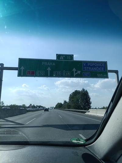 The image shows a view from inside a car on a highway. The sun is high in the sky, indicating it is daytime, and the weather appears to be clear with some clouds. The highway is a multi-lane road with other vehicles, including cars and trucks, visible ahead.
Dominating the scene are two large green highway signs. The left sign directs to "PRAHA" with an upward arrow, and lists road numbers "1", "E50", and "E55" with corresponding arrows. The right sign indicates destinations "V. POPOVICE" and "STRANČICE" with arrows, and shows road number "107" and a distance of "500 m". Above these signs, a smaller sign with "15" and an arrow suggests a highway number or exit.
The overall scene suggests travel on a highway in the Czech Republic, with Všechromy, Czechia, identified as the location. The atmosphere appears to be one of normal traffic flow on a clear day. A green circular sticker with some text, possibly a toll vignette, is visible on the lower right side of the car's dashboard or windshield.