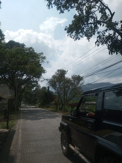 The image depicts a daytime scene in Cibodas, Indonesia, focusing on a paved road flanked by dense foliage under a partly cloudy sky. In the immediate foreground on the right, the front passenger side of a dark-colored, two-door Daihatsu Taft 4x4 vehicle is visible. It features an aggressive off-road tire, a silver-colored metal side step bar, and visible badging on the lower door. A red and white packaged item is visible on the passenger seat through the open window. The paved road recedes into the background, bordered on both sides by numerous tall, green-leafed trees casting shadows. A solid white line marks the left edge of the road. On the far left, a light-colored building with a dark, sloped roof is partially visible, with a dark motorcycle parked in front of it. Overhead utility lines are strung across the upper-right portion of the frame. In the distant background, mountains or hills are visible beneath the cloud-covered sky. No individuals are present in the scene.