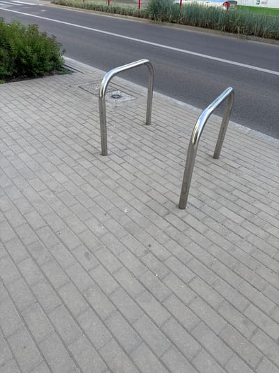The image depicts an outdoor scene on a sidewalk adjacent to a street. The sidewalk is paved with light grey rectangular bricks laid in a herringbone pattern. In the foreground, two U-shaped silver metal bike racks are positioned on the sidewalk, one closer to the viewer and slightly to the right, and the other further away and to the left. The bike racks are anchored into the pavement. Behind the sidewalk, a grey asphalt street is visible with a white line marking a lane. A strip of green foliage, likely ornamental grass or shrubs, separates the street from a building or fence in the background. The overall lighting suggests it is daytime, possibly overcast or late afternoon given the soft shadows. There are no people or vehicles directly visible in the image, though a car and a portion of a building with the text "BKM" are faintly visible in the far background. This scene is likely located in the city of Białystok, Poland, given the visual context and the presence of "BKM" which may refer to a local transportation company.