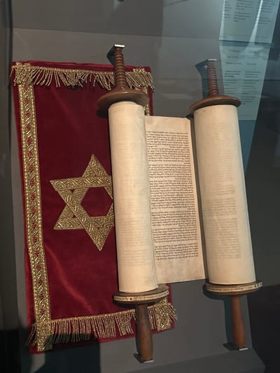 The image depicts a Torah scroll, a sacred text in Judaism, displayed within a museum setting in Bern, Switzerland. The scroll is made of parchment and is unrolled on two wooden rollers, revealing Hebrew script. The Torah is draped over a rich, deep red velvet cloth that is adorned with intricate gold embroidery in the form of a Star of David and decorative borders with fringed tassels. The environment appears to be a dimly lit exhibition space, with a glass partition reflecting some of the surroundings. The focus is on the religious artifact, presented in a manner that emphasizes its historical and cultural significance.