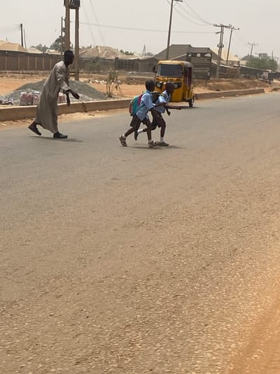 An outdoor scene depicts three individuals crossing an asphalt road under a bright, hazy sky, within Nigeria. An adult male, wearing a light-colored traditional robe and dark shoes, is walking from left to right, with his right arm extended slightly forward and downward. Directly ahead of him, two individuals, appearing to be school-aged boys, are running or jogging in the same direction. Both boys are dressed in matching light blue short-sleeved shirts, plaid shorts, and sandals. The boy on the left carries a light blue backpack. In the midground, a yellow three-wheeled auto-rickshaw is parked on the right shoulder of the road. The background reveals utility poles with overhead wires, multiple low-rise buildings with various roof types, and fences. On the left side of the road, a pile of construction materials is present on a dry, reddish-brown dirt shoulder. The asphalt road surface shows visible wear and dust.