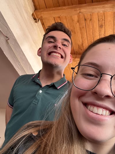 The image displays two young adults indoors. A female individual occupies the right foreground, her right eye and the lower right portion of her face visible. She wears dark-rimmed rectangular glasses and has long, light brown hair. She is smiling broadly, showing her teeth. Behind her, positioned to the left, is a male individual. He has dark, spiky hair and wears a dark green polo shirt with red and white striped trim on the collar and sleeve cuffs. He is also smiling widely, exposing his teeth, with a slightly exaggerated facial expression. The background features a light-colored wall with a prominent horizontal wooden beam and visible red and white wiring in the upper left section. The upper portion of the frame reveals a wooden plank ceiling. The specific location within Dimitrovgrad Municipality, Serbia, is not discernible from the visual information provided.