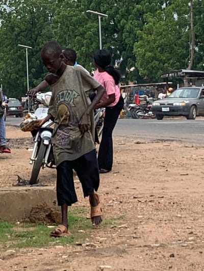 This image captures an outdoor street scene in Shendam, Nigeria, under daylight conditions. The foreground consists of reddish-brown dirt and gravel with sparse patches of green grass, leading to a paved asphalt road in the mid-ground. Dense green foliage from large trees forms the background.
The central figure is a dark-skinned male child or young adolescent with a thin build and short hair. He wears a faded, dirty, light brown t-shirt featuring a dark graphic print (possibly a skull or abstract design), dark blue or black baggy trousers, and light brown open-toed sandals. He is positioned slightly off-center to the left, holding the handlebars of a silver-colored motorcycle with his left hand and a clear plastic container with yellow contents in his right hand. He appears to be walking forward or standing beside the motorcycle.
Behind the central figure, partially obscured, is another dark-skinned individual wearing a light-colored top. To the right of the central figure, a dark-skinned female wearing a pink t-shirt and dark, flared pants is partially visible, oriented away from the viewer.
On the paved road in the mid-ground right, a dark gray sedan (appearing to be a Honda Accord) is present, with its front facing the viewer's left. A white license plate with partially visible characters is affixed to its front bumper. Behind this car, a person in a white t-shirt is visible. Further left, on the same paved road, several motorcycles are parked, and a person in a red top stands near them. Behind these parked motorcycles and people, a makeshift structure with corrugated metal roofing is visible, bearing partial text "HERER OIL & GAS".
In the background, two tall, slender white streetlights with horizontal fixtures stand above the tree line. A utility pole is visible on the far right edge of the frame. The overall impression is a bustling, informal street environment.