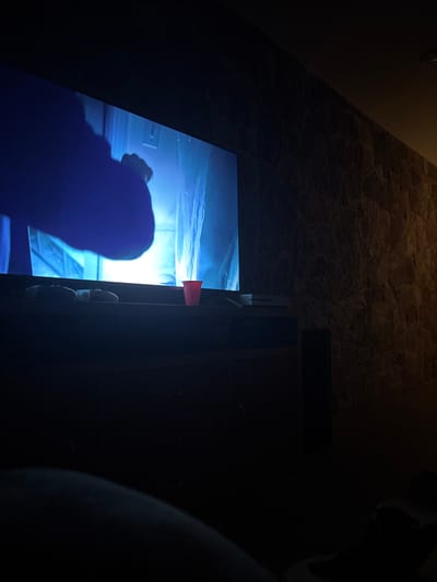 The scene is dimly lit, likely indoors. The primary focus is a glowing screen, possibly a television or computer monitor, which is displaying an image of what appears to be a person, heavily shaded in blue. A portion of the screen is obscured by the edge of a dark, wooden cabinet or piece of furniture. A small, red, cylindrical object is visible near the top edge of the screen and cabinet. The floor and wall are constructed with a stone or rock material.