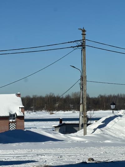 The scene depicts a winter landscape in Shlisselburg, Russia, under a clear, bright blue sky. The ground in the foreground is covered in white snow, displaying vehicle tracks and dark blue shadows cast by unseen objects to the right. To the left, a reddish-brown brick building with a pitched roof heavily covered in snow is partially visible. A window on this building features a vertical white and dark striped pattern directly beneath it.

Dominating the mid-ground is a tall, weathered utility pole, likely concrete or wood, from which numerous black electrical wires extend horizontally across the frame and diagonally downwards. A black street lamp fixture, curved downwards, is attached to this pole. Further to the right, a second, older-style street lamp with a dark, square-shaped lantern atop a pole is visible, surrounded by a large snowdrift.

Behind the utility pole, a smaller, low-slung building with a dark roof and a red brick chimney is partially obscured by snowdrifts. A wide body of water, partially frozen with patches of open, dark blue water and areas of white ice, stretches across the mid-background. Beyond the water, a dense line of bare, brown deciduous trees marks the distant bank, set against the low horizon. No individuals are present or engaged in any discernible actions.