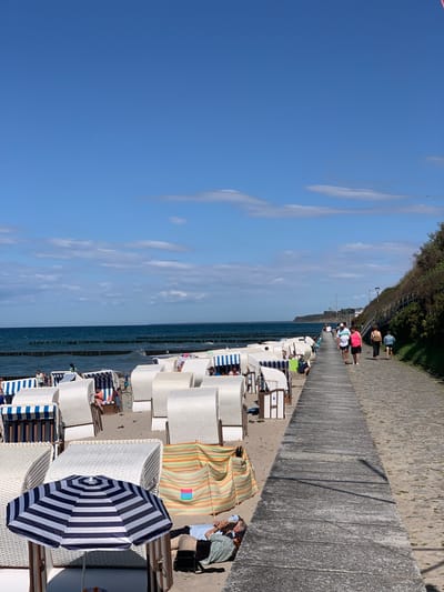 This is a full-frame, eye-level, outdoor, vertical shot of the beach in Nienhagen, Germany. The scene depicts a sunny day with a clear blue sky and some scattered clouds. In the foreground, on the sandy beach, are rows of white wicker beach chairs with blue and white striped canopies. Some people are seated in the chairs, and one person is lying down on the sand under a striped beach umbrella. To the right of the beach chairs is a paved walkway or promenade. Several people are walking on this path, including a couple walking hand-in-hand and another couple walking behind them. The walkway is lined with trees and vegetation on its right side. In the background, the blue ocean stretches to the horizon, with a line of wooden breakwaters visible in the water. There are a few sailboats on the water in the distance. The overall impression is a vibrant and popular beach scene on a pleasant summer day.