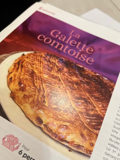 The image displays a close-up photograph of a printed page, likely from a magazine or cookbook. The central focus of the page is a large, appetizing image of a baked good identified as "La Galette comtoise." This pastry is golden-brown with a glossy, textured top surface featuring an intricate, etched pattern resembling a braided or feather-like design. It is depicted resting on a light-colored, possibly white or cream-colored, rectangular platter.

Above the pastry, the title "La Galette comtoise" is printed in prominent pink-hued serif font against a dark purple background. In the upper left corner of the page, the word "DÉGUSTER" is visible in smaller white text. Below the main image, in the bottom left portion of the page, a graphic consisting of two stylized pink figures is present, accompanied by the French text "Pour 6 pers" (For 6 people). To the right of the pastry image, columns of smaller black text are visible, indicating an accompanying article or recipe instructions in French, with fragmented words such as "sud," "site," "vosgie," "vallées," and "Gérard D." The photograph captures the printed page in an indoor setting under ambient lighting, with the background outside the page boundaries appearing dark and out of focus. The physical location of this printed page within the city of Sélestat, France, is not discernible from the visual information provided in the image.