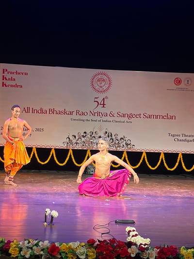 The image shows a performance at Tagore Theatre in Chandigarh, India. The stage is decorated with yellow garlands. Two male dancers are performing on stage. The dancer in the foreground is wearing a pink skirt and a golden belt. He is in a kneeling position, facing the audience. The dancer in the background is wearing a yellow skirt and a golden belt. He is standing, facing the audience. The stage is lit with purple and pink lights. There is a banner behind the dancers with the text "54th All India Bhaskar Rao Nritya & Sangeet Sammelan". There is also a sign in the bottom right corner of the image that says "Tagore Theatre Chandigarh". The coordinates (latitude: 30.733, longitude: 76.79) correspond to the location of Tagore Theatre in Chandigarh, India.