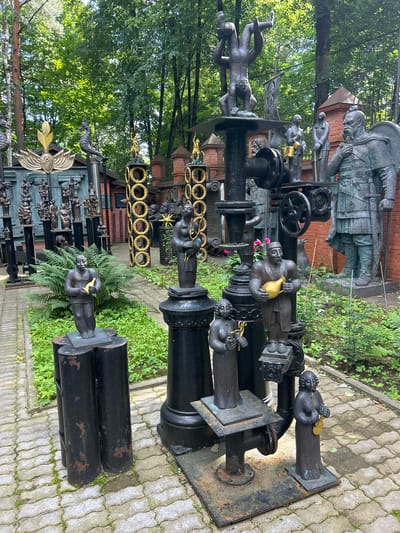 This image shows an outdoor sculpture garden. The sculptures are made of dark metal, and many depict human figures in various poses. Some figures are playing musical instruments, such as a violin and a lute. There are also larger, more imposing figures, some appearing to be knights or warriors. The garden is paved with interlocking stones, and there is lush green vegetation growing around the sculptures. In the background, there are trees and a brick wall. One prominent sculpture in the foreground features a man holding a golden vase. The garden appears to be located in the DSK "Michurinets" residential complex in Russia.