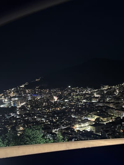 This media file captures a night view of a densely populated area, likely a town or city, illuminated by numerous lights. The image is oriented sideways, with the dark expanse of the night sky occupying the left portion and a lit-up urban landscape filling the right. Buildings of various sizes and shapes are visible, their windows glowing with artificial light, suggesting residential or commercial activity. Amidst the urban sprawl, patches of green foliage indicate the presence of trees. The scene is framed by a thick, light-colored vertical structure on the right edge of the image and a darker, curved structure on the left, possibly indicating a balcony, window frame, or architectural element of the viewing point. The overall impression is that of a vibrant, illuminated cityscape viewed from an elevated position at night. No specific activity or event is clearly discernible, but the widespread illumination points to a lively environment. No text is visible in the image. Given the location context of Košljun, Montenegro, the scene likely depicts a part of this area after dark.