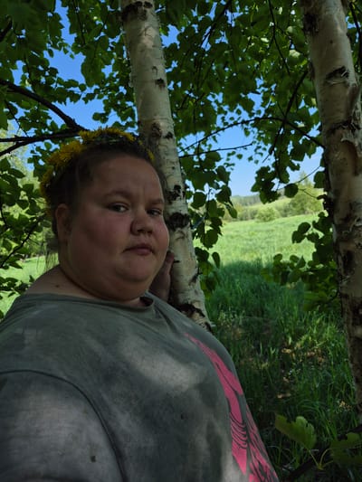 A medium shot, taken outdoors during daylight, shows a woman reclining in a field in the city of Дойная, Russia. She wears a dandelion crown on her head and a gray t-shirt with a pink design. She appears to be leaning against the trunk of a birch tree. A field is visible in the background. The sky, dotted with clouds, is visible through the birch tree branches.