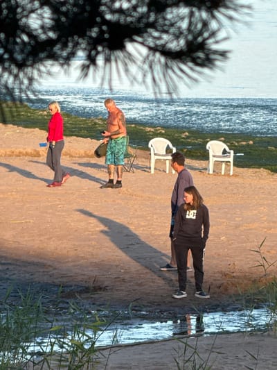 The image depicts a sandy beach area, likely in Shepelevo, Russia, with a body of water in the background. In the foreground, a man with tattoos on his arms and legs, wearing blue patterned shorts and black socks, stands holding a dark cylindrical object. To his left, a woman in a red top and grey pants is walking away from the camera. Further inland, two white plastic chairs are visible. In the mid-ground, a man in a blue long-sleeved shirt and a woman in a black hoodie and black pants are standing. The beach is dotted with sparse green vegetation, and the water is a muted blue with some ripples. The overall lighting suggests it is late afternoon or early evening, casting long shadows across the sand.