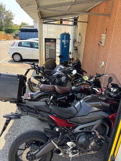 In Fontevivo, Italy, under a covered structure, are three parked motorcycles. The motorcycles are in front of an air compressor. To the left of the motorcycles is a white car. The ground is covered with gravel. The structure has a corrugated metal roof. A brown textured wall is visible to the right. The scene appears to be an outdoor area, possibly a service station or a parking lot.