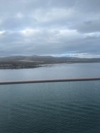 The image captures a panoramic view of a coastline under an overcast sky, observed from an elevated position, likely a vessel, near Stanley, Falkland Islands (Islas Malvinas). In the immediate foreground, a metallic light-grey horizontal railing extends across the lower portion of the frame. Below the railing, a body of choppy, dark blue-green water with visible ripples extends towards the distant landmass. The mid-ground is occupied by a low-lying landmass characterized by undulating, treeless terrain, featuring exposed grey rock formations and sparse, muted green-brown vegetation. No specific structures or signs of human habitation are visible on the land. The background consists of a dense, uniformly grey cloudy sky, indicating heavy overcast conditions with no direct sunlight. No individuals or specific actions are depicted.