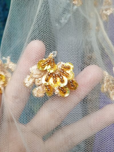 A close-up, high-angle shot captures a translucent, light-beige netting fabric adorned with intricate golden and amber embellishments. A human hand, likely an adult's, is positioned beneath the sheer fabric, with fingers spread, creating a blurred silhouette visible through the mesh. The skin tone of the hand appears light.
The fabric is a fine mesh or tulle, featuring a distinct, repeating hexagonal pattern of small holes. Embroidered onto this netting are clusters of embellishments, primarily golden. These consist of small, circular, iridescent gold sequins, which form the base of floral or leaf-like patterns. Tiny, amber-colored seed beads are stitched on top of the sequins, often arranged in rows or small clusters, adding texture and dimension. Some areas also show light-beige or cream-colored threadwork that blends with the fabric.
The background is softly blurred, revealing indistinct patches of dark teal, muted green, and purple colors visible through the sheer material and around the edges of the frame. The lighting is soft and even, highlighting the reflective quality of the sequins and the delicate structure of the netting. No text is visible in the image. The scene focuses on detailed textile craftsmanship, possibly for apparel or decoration, originating from Houaykhang, Laos.
