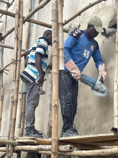 Two men are working on a construction site in Okinano, Nigeria. The man on the left is wearing a striped t-shirt and gray pants, and is holding a hammer. The man on the right is wearing a blue long-sleeved shirt with the number "48" on it and black pants. He is wearing a green cap and protective gloves, and is operating an angle grinder. Both men are standing on a wooden platform supported by bamboo scaffolding, with a concrete wall in the background. The scene depicts an active construction environment.