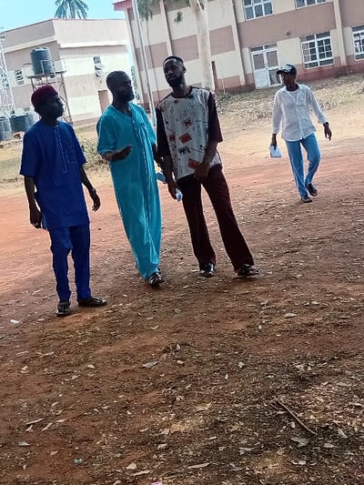 Four men stand on a dirt ground. The man in front wears a blue traditional outfit and a maroon cap. Behind him, another man is dressed in a turquoise robe. The third man dons a graphic t-shirt and brown pants. The last man is dressed in a white shirt and blue jeans, also wearing a cap. The background shows a building with multiple windows and some trees.