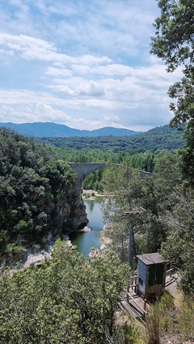 An outdoor, eye-level, vertical shot captures a stone bridge spanning a river in Tortellà, Spain. The bridge, with its arched structure, is a prominent feature in the mid-ground. The river flows through a rocky gorge, flanked by lush green trees and vegetation on either side. In the background, rolling hills covered in dense forest recede into the distance, under a sky dotted with white clouds. In the foreground, to the right of the frame, a small, gray, rectangular building with a dark door sits on a concrete base, surrounded by a wooden railing. A metal pole with some equipment attached stands beside the building. The overall scene depicts a natural landscape with a historical structure.