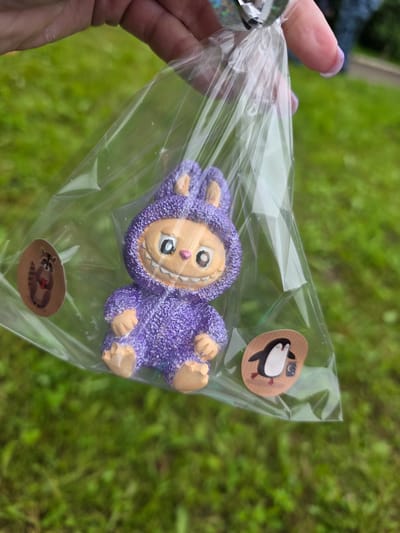 A person's hand is holding a clear plastic bag containing a small, purple, fuzzy figurine. The figurine resembles a cartoonish creature with a wide smile, big eyes, and a pink nose. It has tan-colored feet and hands, and its body is covered in sparkly purple fuzz. There are two small, circular stickers on the outside of the plastic bag. One sticker, near the top right, features a cartoon penguin. The other sticker, near the bottom, shows a cartoon raccoon holding a red heart. The background is out of focus and appears to be green grass, suggesting an outdoor setting. The lighting is bright, indicating it is daytime. The hand holding the bag has fingernails painted with a light purple color. The context provided indicates the location is Старые Быги, Russia.