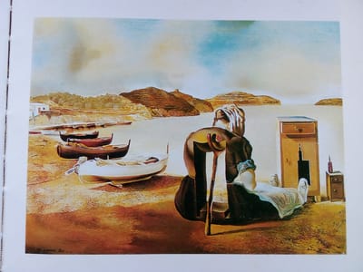 The image depicts a surreal landscape with a body of water and a rocky coastline. In the foreground, several boats are beached on a sandy shore. A figure, appearing to be an elderly person, is seated with a crutch. To the right, a drawer unit and a bottle are visible. The sky is rendered in soft, muted tones. There is no indication of the city of Zhmerynka, Ukraine, within the image.