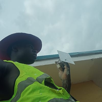 A construction worker in Gana Daji, Nigeria, wears a red hard hat and a tattered lime green and gray safety vest. He is using a rectangular trowel to apply plaster or mortar to a building's exterior. His hands are covered in the white material. The sky is overcast.