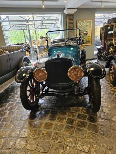 The image showcases a vintage car, possibly a Model T Ford, displayed inside a building, likely a museum, in Архангельское, Russia. The car is teal in color with black fenders and prominent brass headlights. It's positioned on a cobblestone floor. In the background, a large window provides a view of greenery, and another vintage vehicle can be seen through the window, along with a painting on the wall. The overall setting evokes a historical display or exhibit.