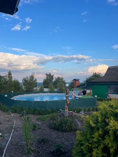 The image depicts an outdoor scene at dusk. Two people are visible in a swimming pool, one standing and the other sitting. The pool is oval-shaped and appears to be above ground. A decorative statue of a woman holding a sphere stands near the edge of the pool. The setting includes a building with a dark roof, lush green trees and bushes, and a cloudy sky with hues of orange and pink, indicating sunset. The ground is covered with dark soil and some rocks. The overall atmosphere suggests a relaxed, leisurely evening. No text is visible in the image.
