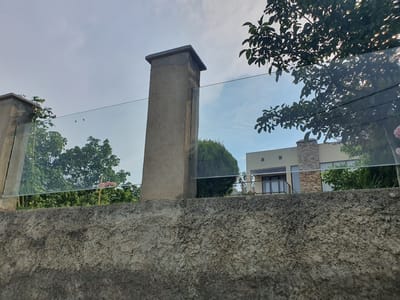 The image captures a view of a modern building and surrounding greenery in Mukhatskaro, Georgia. In the foreground, a textured concrete wall runs horizontally across the frame. Above this wall, a clear glass railing, supported by concrete pillars, divides the scene. The taller pillar, located to the left of center, has a square cap. Beyond the glass railing, a two-story building with beige siding and stone accents is visible. A balcony with glass railings is on the second floor, and large windows are on both levels. Lush green trees and vegetation surround the building. The sky above is a light blue with scattered clouds. The overall impression is one of a well-maintained residential or commercial property.