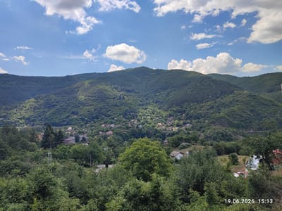 This media file captures a scenic view of a village nestled within a lush, green mountainous landscape under a bright, partly cloudy sky. The scene is dominated by the verdant slopes of the mountains, densely covered with trees. A small village is visible, with clusters of houses scattered among the trees on the hillsides. In the foreground, there is a dense canopy of green foliage, with the tops of trees and bushes extending towards the viewer. Power lines and a green metal pole are also visible in the mid-ground, suggesting the presence of infrastructure. On the far right, a dilapidated structure with a red, corrugated metal roof is partially visible, with some vehicles, including what appears to be a white car and a red car, parked nearby. The overall impression is one of a peaceful, rural setting in a mountainous region. The time stamp "19.06.2026 15:13" indicates the photograph was taken on June 19, 2026, at 3:13 PM, suggesting it is mid-afternoon on a sunny day, consistent with the bright lighting and scattered clouds. The location context places this scene in Tompsan, Bulgaria. No people are visible in the image.