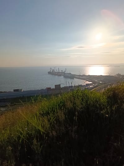 An elevated, wide-angle, natural light photograph captures a coastal industrial area in Korsakov, Russia, under a bright, partly cloudy sky at sunrise or sunset. The foreground is dominated by a slope covered in dry and green tall grass, with some lower, tilled fields visible beneath.
The middle ground reveals a coastal port with several large cargo ships docked. Cranes, characteristic of port operations, are positioned on the ships and the shore. A long, low-rise industrial building complex with multiple rooflines stretches along the waterfront. Adjacent to this, railway tracks and various containers are visible. The sea, reflecting the bright sun, occupies the majority of the image, extending to the horizon.
In the distance, several more ships are visible on the water, and a faint, hazy landmass can be seen on the far horizon. The sun, a bright orb, is positioned in the upper right quadrant of the sky, casting a warm glow and lens flare across the scene. No people are discernible in the image.
