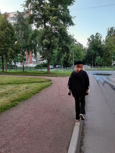 A person with long, blonde hair, wearing a black beret, black hoodie with white flame-like accents on the sleeves, and black pants walks along the curb of a paved road. The person is viewed from behind and is positioned in the lower right quadrant of the frame. To the left of the road is a gravel path, and further to the left is a patch of green grass. In the background, a multi-story building with red and white siding is visible, partially obscured by numerous trees with green foliage. The sky above is a pale blue. The scene appears to be in a residential area, possibly in the city of Kolpino, Russia, given the architectural style and surrounding vegetation. There are cars parked on the road in the distance. The road surface is wet, indicating recent rain or cleaning.