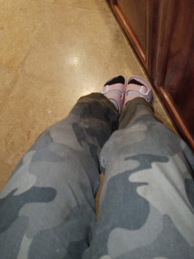 The image displays a downward perspective of a person's lower extremities within an interior setting in Madinaty, Egypt. The individual is wearing grey and dark grey camouflage-patterned pants. Their feet are clad in black socks and light pink, two-strap sandals featuring white buckles. The subject is positioned on a light tan-colored floor, possibly tile or linoleum. To the right of the feet, a dark brown wooden vertical surface, exhibiting visible horizontal grooves, is present, likely a piece of furniture or an architectural element like a baseboard or wall panel.