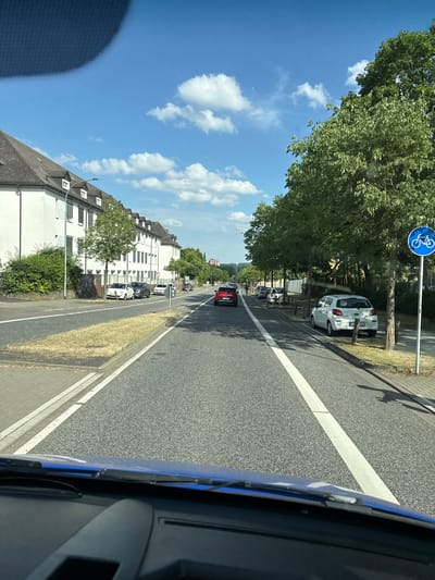 The image depicts a street scene in Giessen, Germany, on a sunny day. A car's perspective from the driver's seat shows a road with a white lane marker, a sidewalk, and buildings lining the street. Trees are present along the road, casting shadows on the pavement. Several cars are visible, parked along the side of the road and in the lane of traffic. One car, a white hatchback, is parked at an unusual angle, leaning against a wooden post, with a license plate that reads "GI.T 1090". A blue sign with a white bicycle symbol indicates a bike lane. The buildings are multi-story structures with white facades and tiled roofs. The sky is blue with scattered white clouds. The scene suggests a typical urban environment during daytime. There are no people visible in the frame.
