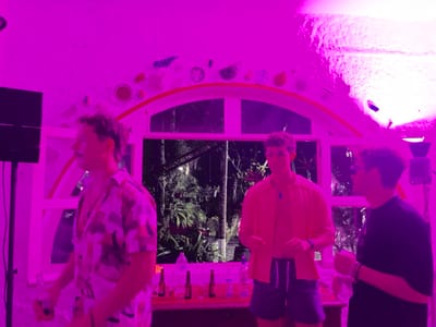 The scene is an indoor space in Fortín de las Flores, Mexico, illuminated predominantly by an intense magenta light. The room features textured white walls and ceiling. A large, arched window with white frames and multiple panes offers a view of dark, dense tropical foliage outside. Above the arch, decorative plates or ceramic objects are affixed to the wall.
Three adult males are present. On the far left, a man with a mustache and wavy dark hair, wearing a patterned short-sleeved shirt and light-colored bottoms, stands in profile, holding a small red object in his left hand. In the center, a man with short brown hair and glasses wears an unbuttoned peach-colored short-sleeved shirt over a darker garment, paired with striped shorts, and a dark pendant around his neck; his hands are loosely clasped in front. On the right, a third man, partially visible, wears a dark short-sleeved shirt and holds a small object in his left hand, displaying a wristband.
A counter or bar runs along the base of the window, holding multiple bottles, including what appear to be liquor bottles. A black speaker is positioned on the left, and a pink-emitting light fixture is visible on the upper right side of the room. The individuals are standing, appearing to be at a social gathering.