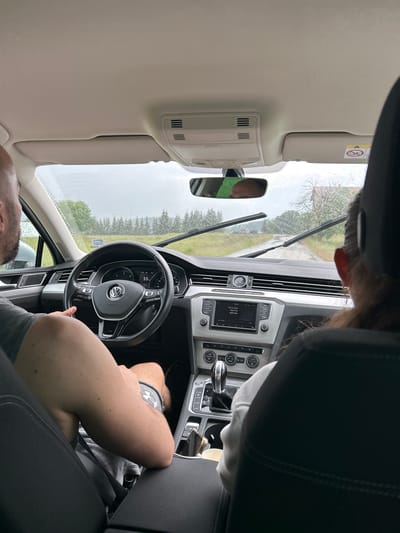 The image shows the interior of a car, likely a Volkswagen, viewed from the back seat looking forward. The scene is dominated by the car's interior, including the dashboard, steering wheel, and parts of the seats. Two individuals are visible. One person, likely the driver, is in the foreground, with their arm resting on the center console and their hand on the steering wheel. Another person is visible in the rearview mirror, reflected from what appears to be the passenger seat.
The car is on a road, as indicated by the windshield view showing trees and a cloudy sky. The weather appears to be overcast and possibly rainy, as streaks are visible on the windshield. The location is identified as Cherna Mesta, Bulgaria.
Notable details include the car's infotainment system displaying song information ("Procesal Jam," "High Ride," "High Ride"). The dashboard shows a speed of 55. A warning label with a "no entry" symbol is visible on the overhead console. The lighting suggests it is daytime, but the cloudy weather creates a diffused light. The overall mood is relaxed, with the individuals seemingly on a journey.