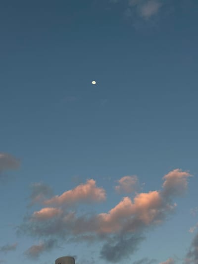 The image displays an outdoor scene in Beit Yitshak Sha'ar Hefer, Israel, focusing primarily on the sky during either daylight or twilight hours. A bright, nearly full white moon is visible in the upper-center portion of the frame against a clear, deep blue sky. Below the moon, a band of scattered cumulus clouds occupies the lower third of the sky, exhibiting a warm, peachy-orange illumination on their undersides, consistent with sunrise or sunset lighting. A partial view of a light grey, textured cylindrical structure is visible at the extreme bottom-center edge of the frame. No individuals, discernible actions, or interactions are present.